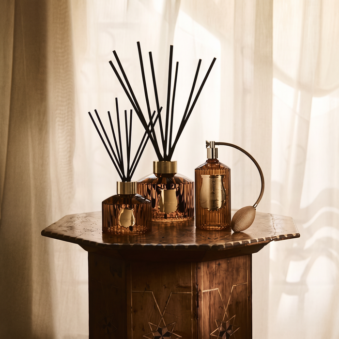 Three amber glass reed diffusers with black reeds and a matching amber atomizer bottle arranged on a rustic wooden table in front of soft cream curtains.