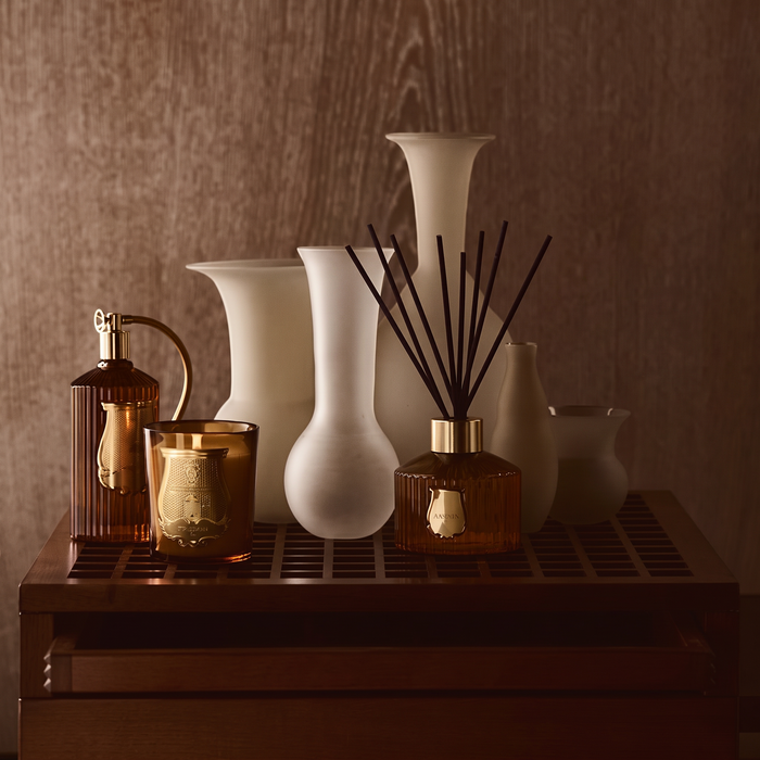 A styled collection of home decor items on a dark wooden tray, including matte white ceramic vases, amber glass bottles, and a reed diffuser, set against a warm brown wood-paneled background.