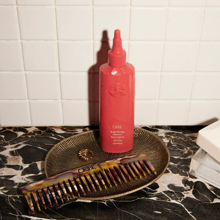 Oribe Bright Blonde Radiance & Repair Treatment bottle with a tortoiseshell comb and gold ring on a marble surface.