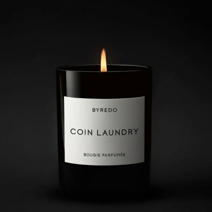 Shop Rescue Spa Byredo Coin Laundry candle in a black glass jar with a lit wick. Floral scent with notes of aldehyde, pink pepper, rose, neroli, peony, violet, woods, musk, and sandalwood.
