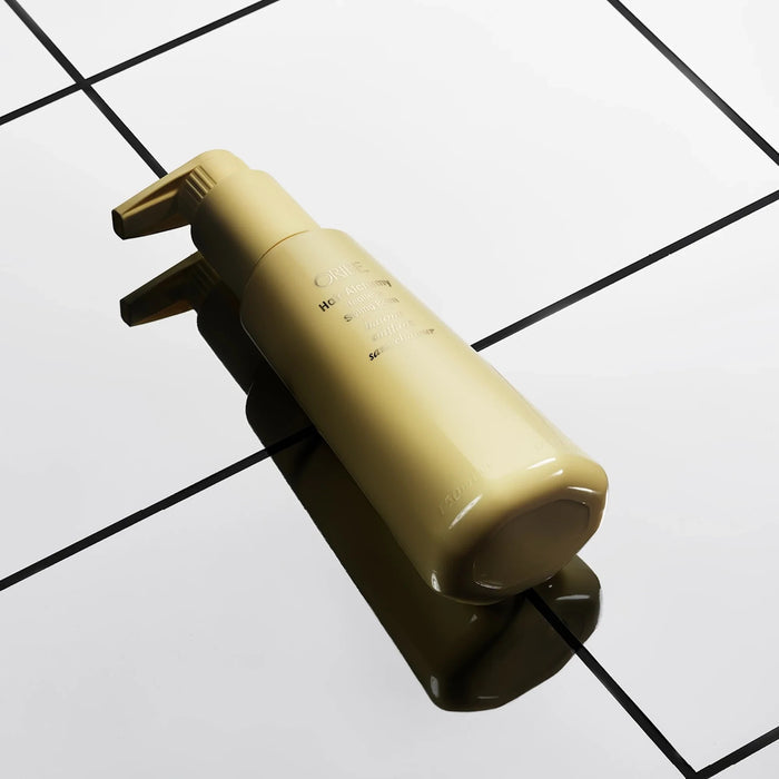 Shop Rescue Spa Oribe Hair Alchemy Heatless Styling Balm in a gold pump bottle on a reflective tiled surface.