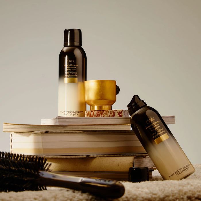 Shop Rescue Spa Oribe Imperial Blowout styling creme bottles on a stack of books with a gold cup and hair brush