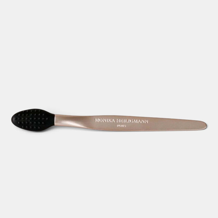 A metallic rose gold massage tool with a black silicone head, featuring the brand name 'MONIKA HELLMANN' engraved on the handle, set against a light gray background.
