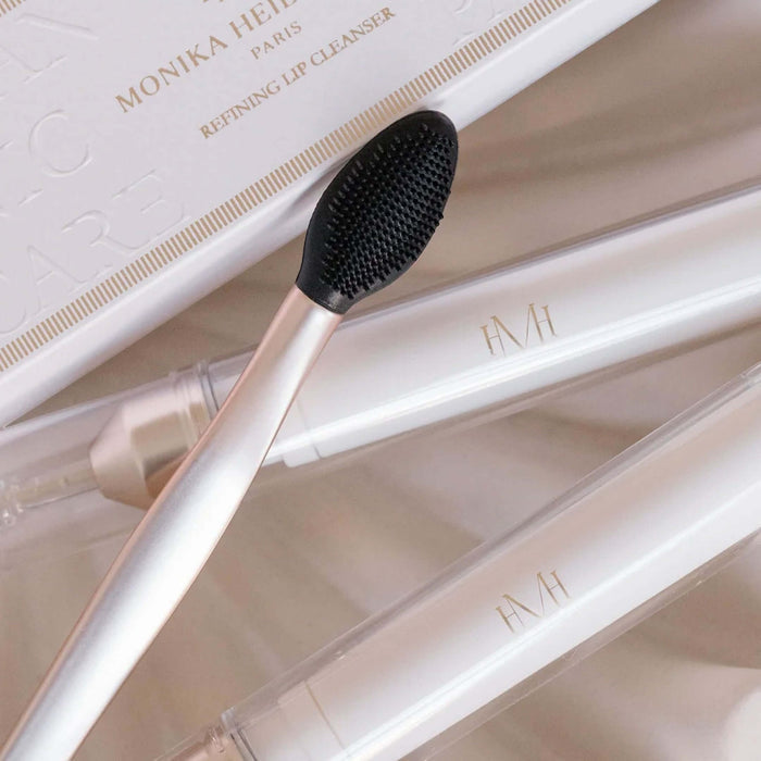 A close-up of a lip cleansing brush with a black bristle head and pale pink handle, resting on a white box labeled Monika Heide Paris Refining Lip Cleanser. Two white cosmetic tubes with gold 'FMI' branding are also visible in a soft, neutral setting.