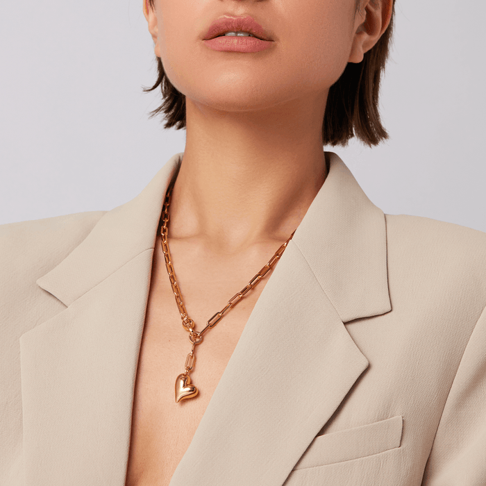 A close-up of a model wearing a gold heart pendant necklace with a chunky chain, styled with a beige blazer.