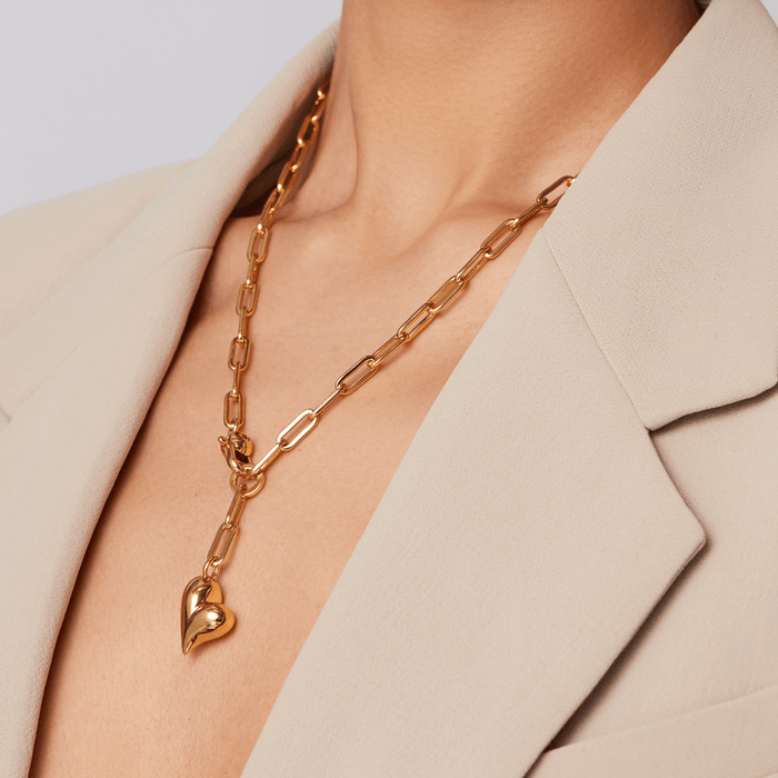 A close-up of a model wearing a gold chain necklace with a heart-shaped pendant, styled with a beige blazer.