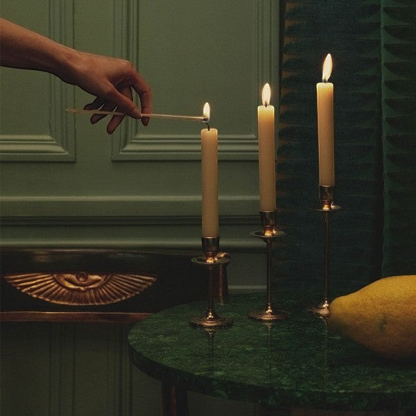 Hand lighting Trudon gold-plated candlesticks with Madeleine taper candles in a dimly lit room, glowing candle flames on tables