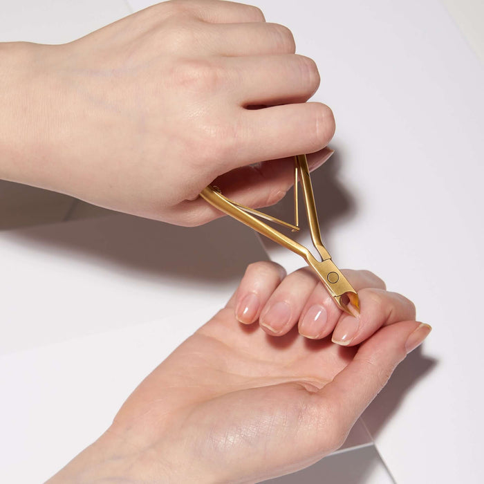 A close-up of hands using a gold metal cuticle pusher or manicure tool on a fingernail against a clean white background.