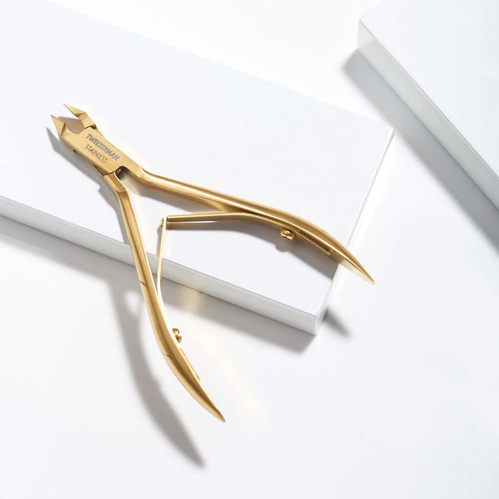 Gold-colored nail clippers with engraved branding, resting on a clean white geometric surface.